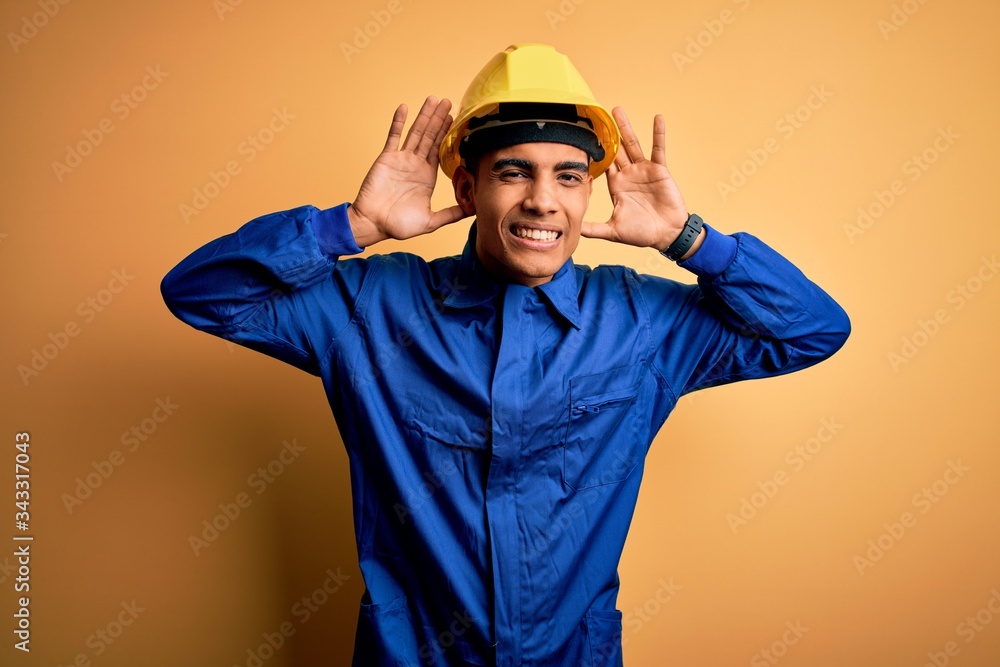 Young handsome african american worker man wearing blue uniform and security helmet Trying to hear both hands on ear gesture, curious for gossip. Hearing problem, deaf