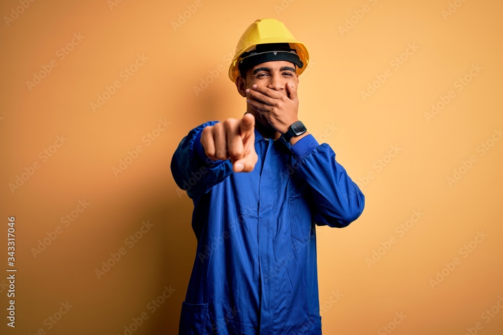 Young handsome african american worker man wearing blue uniform and security helmet laughing at you, pointing finger to the camera with hand over mouth, shame expression