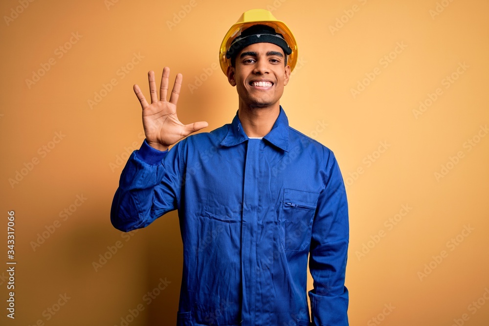 Young handsome african american worker man wearing blue uniform and security helmet showing and pointing up with fingers number five while smiling confident and happy.
