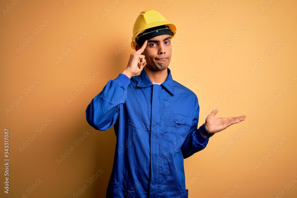 Young handsome african american worker man wearing blue uniform and security helmet confused and annoyed with open palm showing copy space and pointing finger to forehead. Think about it. Young handsome african american worker man wearing blue uniform and security helmet confused and annoyed with open palm showing copy space and pointing finger to forehead. Think about it.