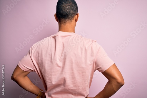 Handsome african american man wearing casual t-shirt and glasses over pink background standing backwards looking away with arms on body