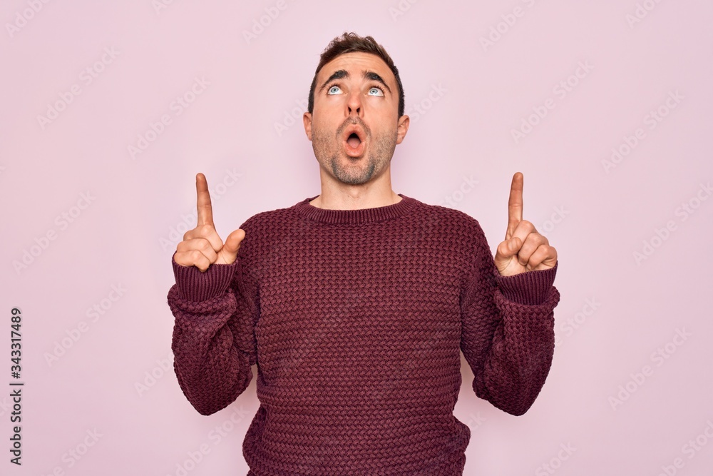 Young handsome man with blue eyes wearing casual sweater standing over pink background amazed and surprised looking up and pointing with fingers and raised arms. Young handsome man with blue eyes wearing casual sweater standing over pink background amazed and surprised looking up and pointing with fingers and raised arms.