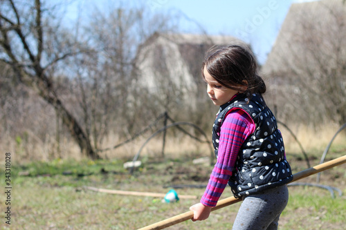 a little white dark-haired white girl with a ponytail outdoors holds a long wooden stick in her hands. Preschooler helps parents in the spring garden