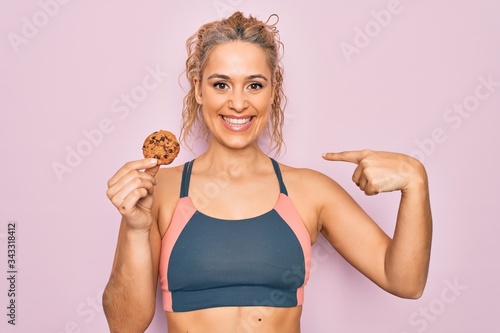 Young beautiful blonde sporty woman doing sport holding cookie over isolated pink background pointing finger to one self smiling happy and proud