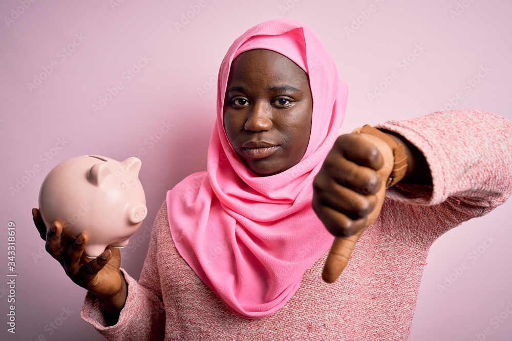 Young african american plus size woman wearing muslim hijab holding pink piggy bank with angry face, negative sign showing dislike with thumbs down, rejection concept Young african american plus size woman wearing muslim hijab holding pink piggy bank with angry face, negative sign showing dislike with thumbs down, rejection concept