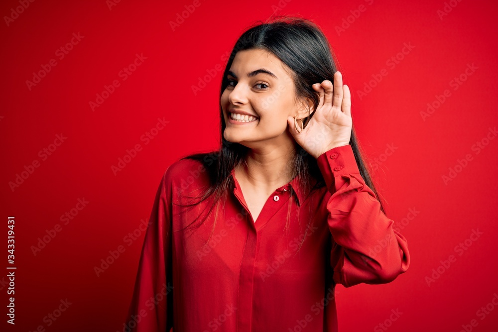 Young beautiful brunette woman wearing casual shirt standing over red background smiling with hand over ear listening an hearing to rumor or gossip. Deafness concept. Young beautiful brunette woman wearing casual shirt standing over red background smiling with hand over ear listening an hearing to rumor or gossip. Deafness concept.