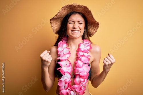 Young beautiful woman with blue eyes on vacation wearing bikini and hawaiian lei excited for success with arms raised and eyes closed celebrating victory smiling. Winner concept.