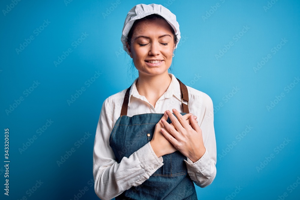 Young beautiful baker woman with blue eyes wearing apron and cap over blue background smiling with hands on chest with closed eyes and grateful gesture on face. Health concept. Young beautiful baker woman with blue eyes wearing apron and cap over blue background smiling with hands on chest with closed eyes and grateful gesture on face. Health concept.