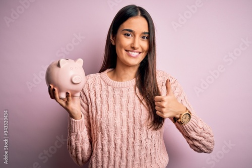 Young beautiful brunette woman holding piggy bank saving money for retirement happy with big smile doing ok sign, thumb up with fingers, excellent sign