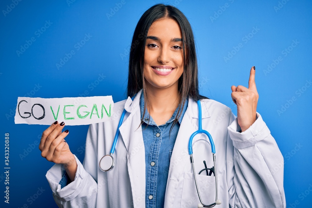 Young beautiful brunette doctor woman wearing coat holding paper with go vegan message surprised with an idea or question pointing finger with happy face, number one Young beautiful brunette doctor woman wearing coat holding paper with go vegan message surprised with an idea or question pointing finger with happy face, number one