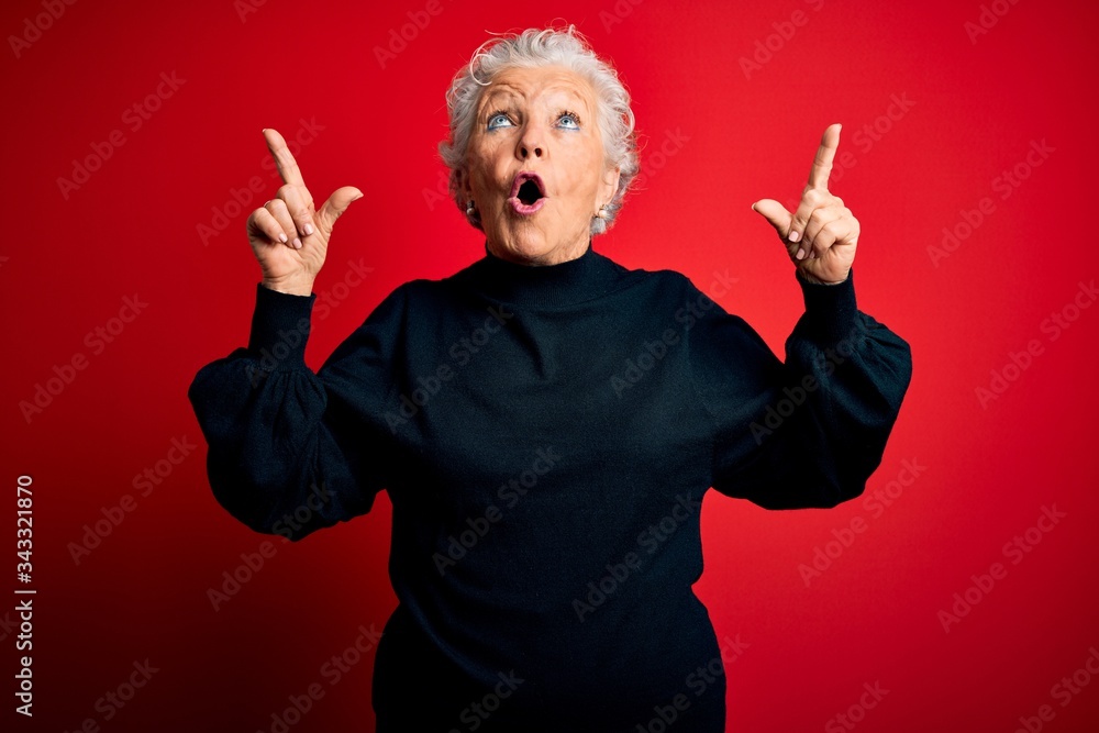 Senior beautiful woman wearing casual sweater standing over isolated red background amazed and surprised looking up and pointing with fingers and raised arms.