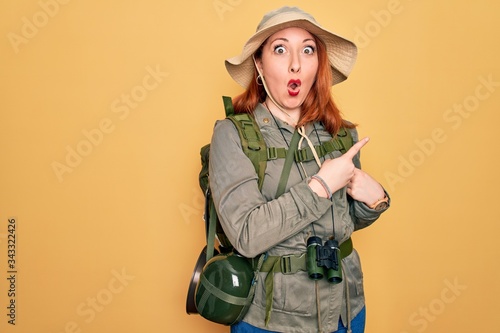 Young redhead backpacker woman hiking wearing backpack and hat over yellow background Surprised pointing with finger to the side, open mouth amazed expression.