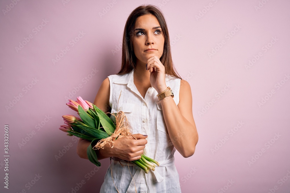 Young beautiful brunette woman holding bouquet of tulips flowers over pink background with hand on chin thinking about question, pensive expression. Smiling with thoughtful face. Doubt concept. Young beautiful brunette woman holding bouquet of tulips flowers over pink background with hand on chin thinking about question, pensive expression. Smiling with thoughtful face. Doubt concept.