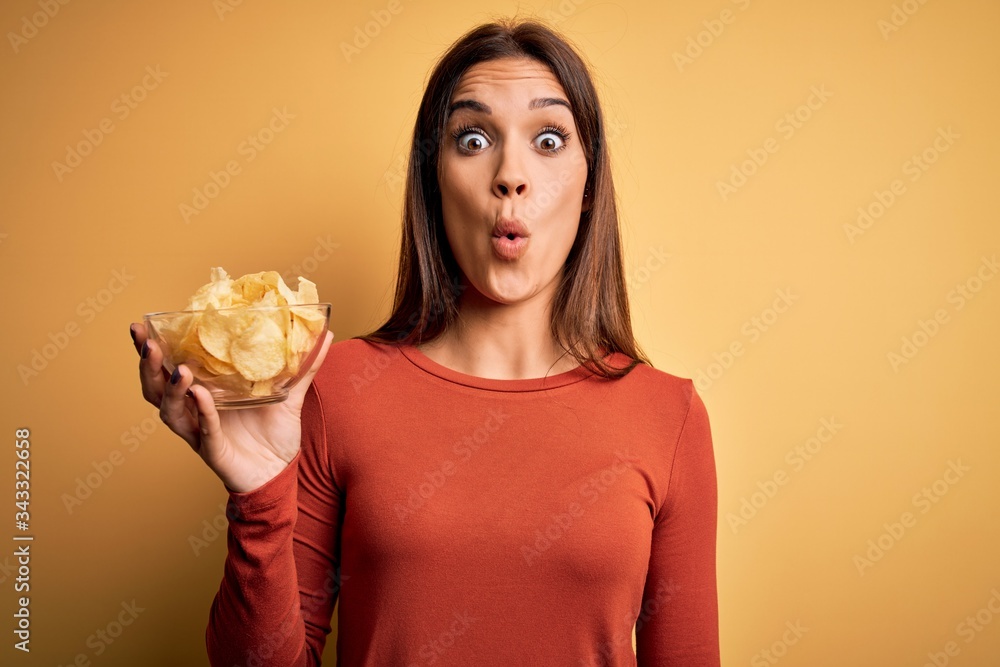 Young beautiful brunette woman holding bowl with chips potatoes over yellow background scared in shock with a surprise face, afraid and excited with fear expression Young beautiful brunette woman holding bowl with chips potatoes over yellow background scared in shock with a surprise face, afraid and excited with fear expression