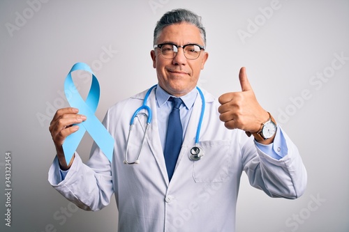 Middle age handsome grey-haired doctor man wearing coat holding blue cancer ribbon happy with big smile doing ok sign, thumb up with fingers, excellent sign