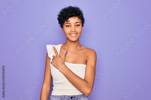Young beautiful african american afro woman wearing casual t-shirt over purple background cheerful with a smile on face pointing with hand and finger up to the side with happy and natural expression