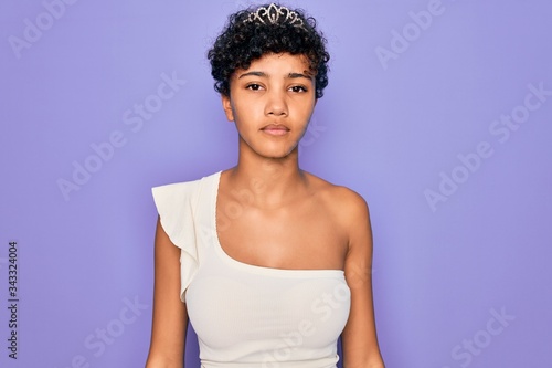 Young beautiful african american afro woman wearing tiara crown over purple background with serious expression on face. Simple and natural looking at the camera.