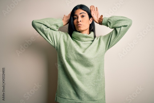 Young beautiful hispanic woman wearing green winter sweater over isolated background Doing bunny ears gesture with hands palms looking cynical and skeptical. Easter rabbit concept. © Krakenimages.com