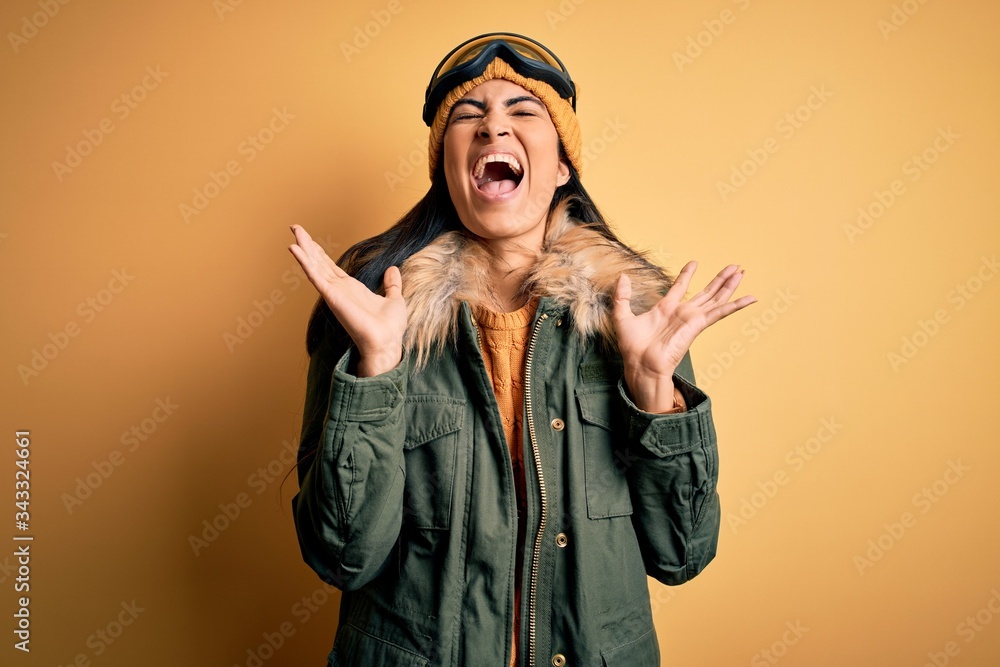 Young beautiful hispanic woman wearing ski glasses and coat for winter weather celebrating mad and crazy for success with arms raised and closed eyes screaming excited. Winner concept Young beautiful hispanic woman wearing ski glasses and coat for winter weather celebrating mad and crazy for success with arms raised and closed eyes screaming excited. Winner concept