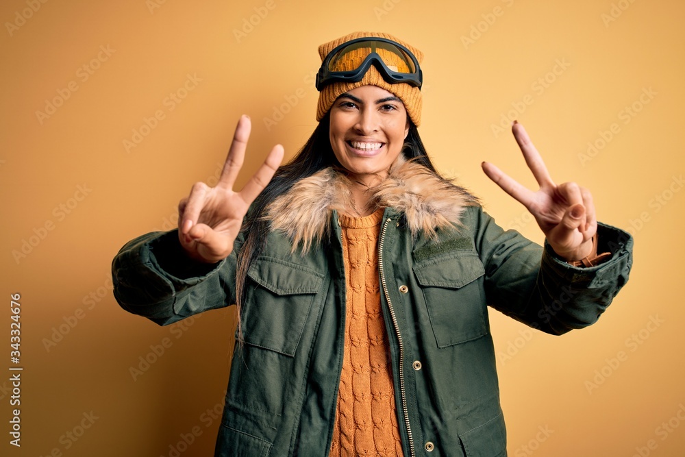 Young beautiful hispanic woman wearing ski glasses and coat for winter weather smiling with tongue out showing fingers of both hands doing victory sign. Number two.