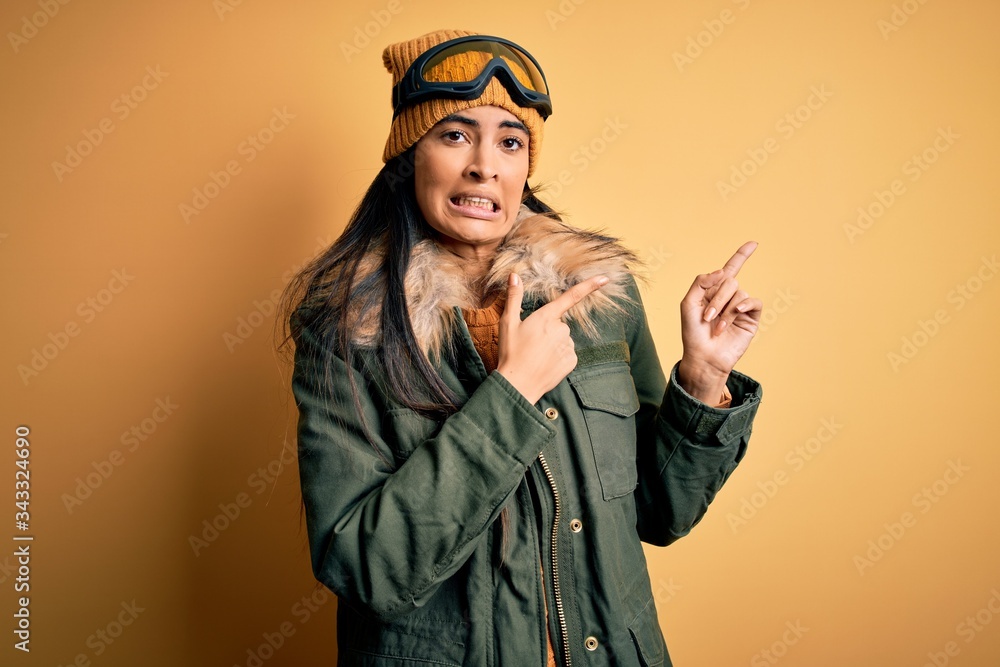 Young beautiful hispanic woman wearing ski glasses and coat for winter weather Pointing aside worried and nervous with both hands, concerned and surprised expression Young beautiful hispanic woman wearing ski glasses and coat for winter weather Pointing aside worried and nervous with both hands, concerned and surprised expression