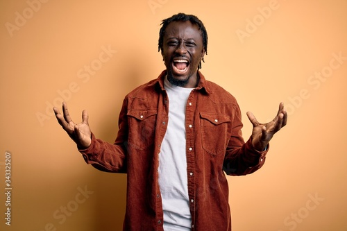 Young handsome african american man wearing casual jacket standing over yellow background crazy and mad shouting and yelling with aggressive expression and arms raised. Frustration concept.