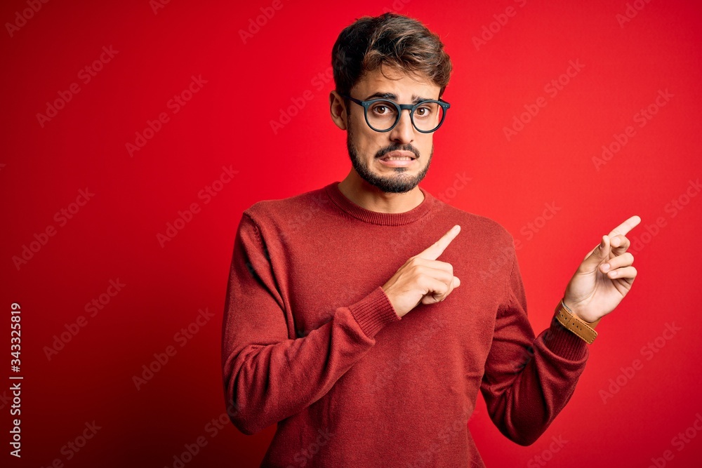 Young handsome man with beard wearing glasses and sweater standing over red background Pointing aside worried and nervous with both hands, concerned and surprised expression Young handsome man with beard wearing glasses and sweater standing over red background Pointing aside worried and nervous with both hands, concerned and surprised expression