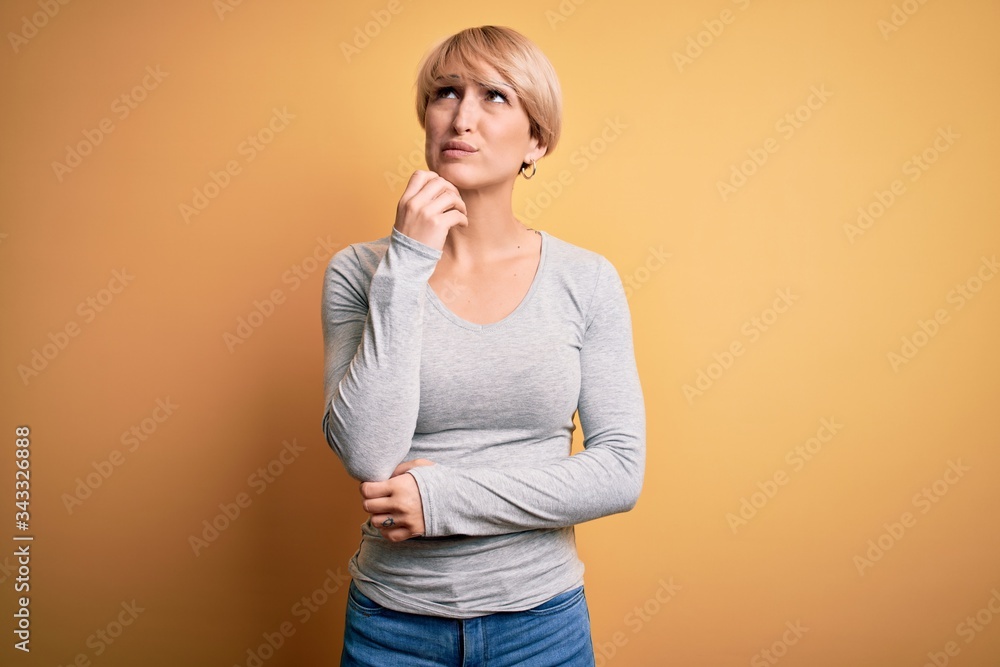 Young blonde woman with modern short hair standing over yellow background with hand on chin thinking about question, pensive expression. Smiling with thoughtful face. Doubt concept. Young blonde woman with modern short hair standing over yellow background with hand on chin thinking about question, pensive expression. Smiling with thoughtful face. Doubt concept.