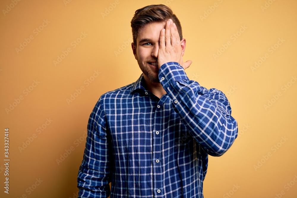 Young blond businessman with beard and blue eyes wearing shirt over yellow background covering one eye with hand, confident smile on face and surprise emotion.
