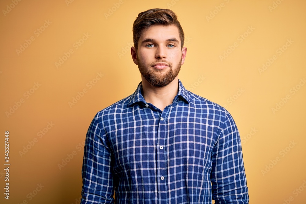 Young blond businessman with beard and blue eyes wearing shirt over yellow background Relaxed with serious expression on face. Simple and natural looking at the camera.