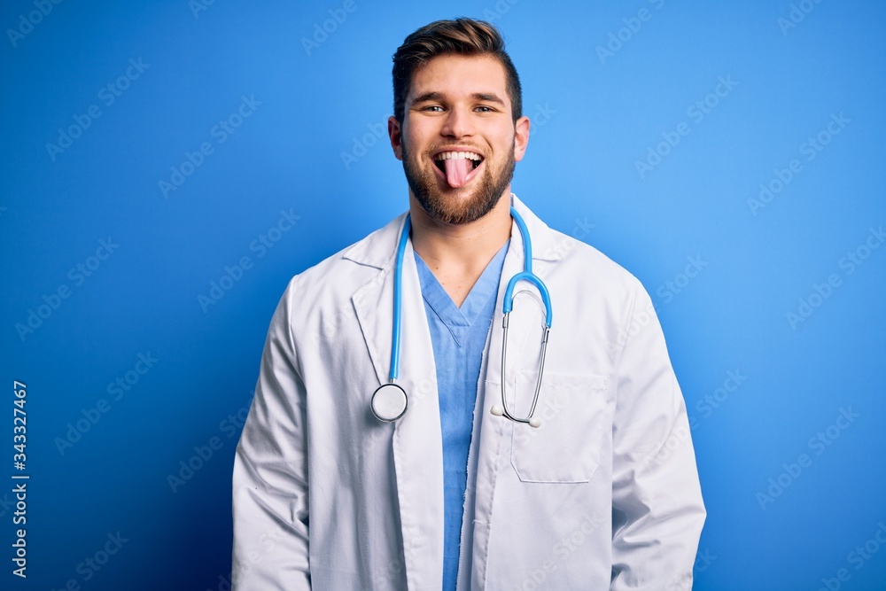 Young blond doctor man with beard and blue eyes wearing white coat and stethoscope sticking tongue out happy with funny expression. Emotion concept. Young blond doctor man with beard and blue eyes wearing white coat and stethoscope sticking tongue out happy with funny expression. Emotion concept.