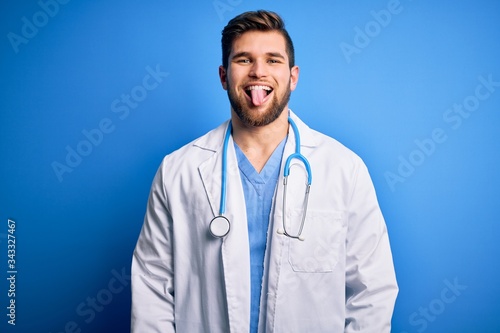 Young blond doctor man with beard and blue eyes wearing white coat and stethoscope sticking tongue out happy with funny expression. Emotion concept.