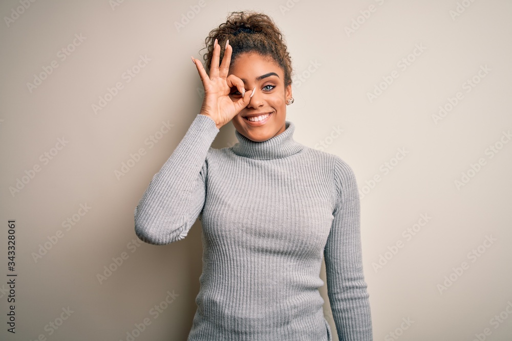 Beautiful african american girl wearing turtleneck sweater standing over white background doing ok gesture with hand smiling, eye looking through fingers with happy face. Beautiful african american girl wearing turtleneck sweater standing over white background doing ok gesture with hand smiling, eye looking through fingers with happy face.