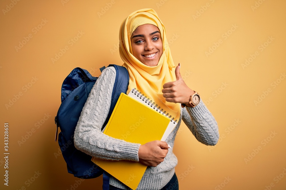Young african american student girl wearing muslim hijab and backpack holding notebook happy with big smile doing ok sign, thumb up with fingers, excellent sign Young african american student girl wearing muslim hijab and backpack holding notebook happy with big smile doing ok sign, thumb up with fingers, excellent sign