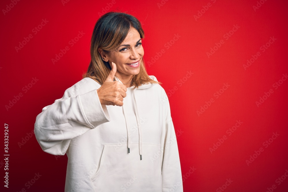 Middle age beautiful sportswoman wearing casual sweatshirt over isolated red background doing happy thumbs up gesture with hand. Approving expression looking at the camera showing success. Middle age beautiful sportswoman wearing casual sweatshirt over isolated red background doing happy thumbs up gesture with hand. Approving expression looking at the camera showing success.