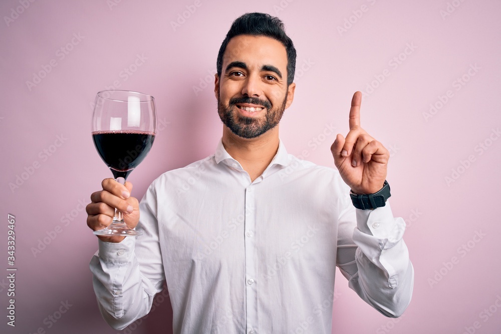 Young handsome man with beard drinking glass of red wine over isolated pink background surprised with an idea or question pointing finger with happy face, number one Young handsome man with beard drinking glass of red wine over isolated pink background surprised with an idea or question pointing finger with happy face, number one