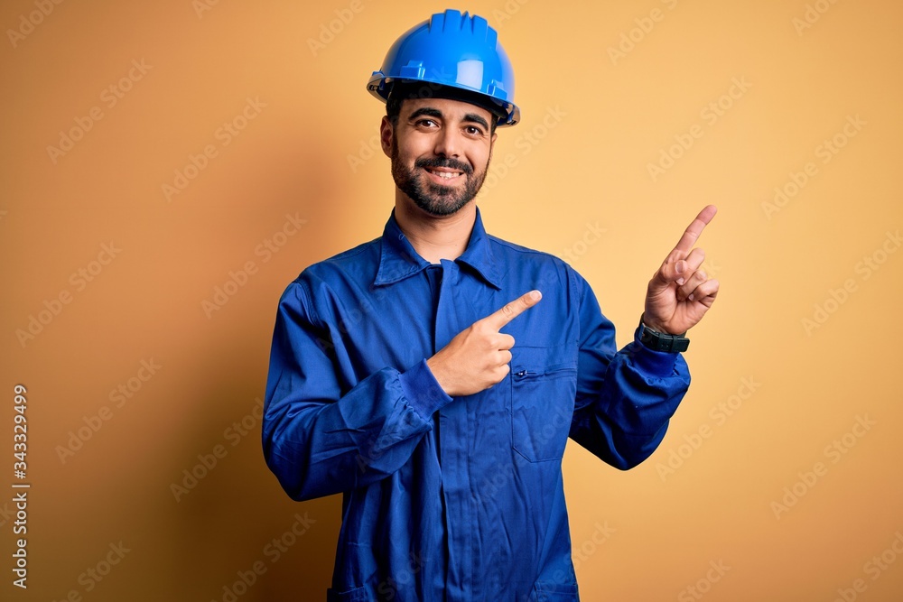 Mechanic man with beard wearing blue uniform and safety helmet over yellow background smiling and looking at the camera pointing with two hands and fingers to the side.