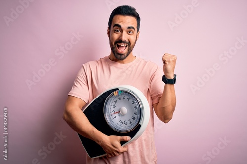 Young handsome slim sportsman with beard holding scale over isolated pink background screaming proud and celebrating victory and success very excited  cheering emotion