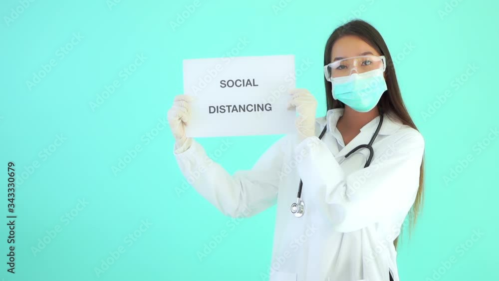 Static shot of a female Asian doctor wearing a coat, stethoscope, surgical mask, and protective eyewear in front of a turquoise background holding a social distancing sign and giving a thumbs up Static shot of a female Asian doctor wearing a coat, stethoscope, surgical mask, and protective eyewear in front of a turquoise background holding a social distancing sign and giving a thumbs up