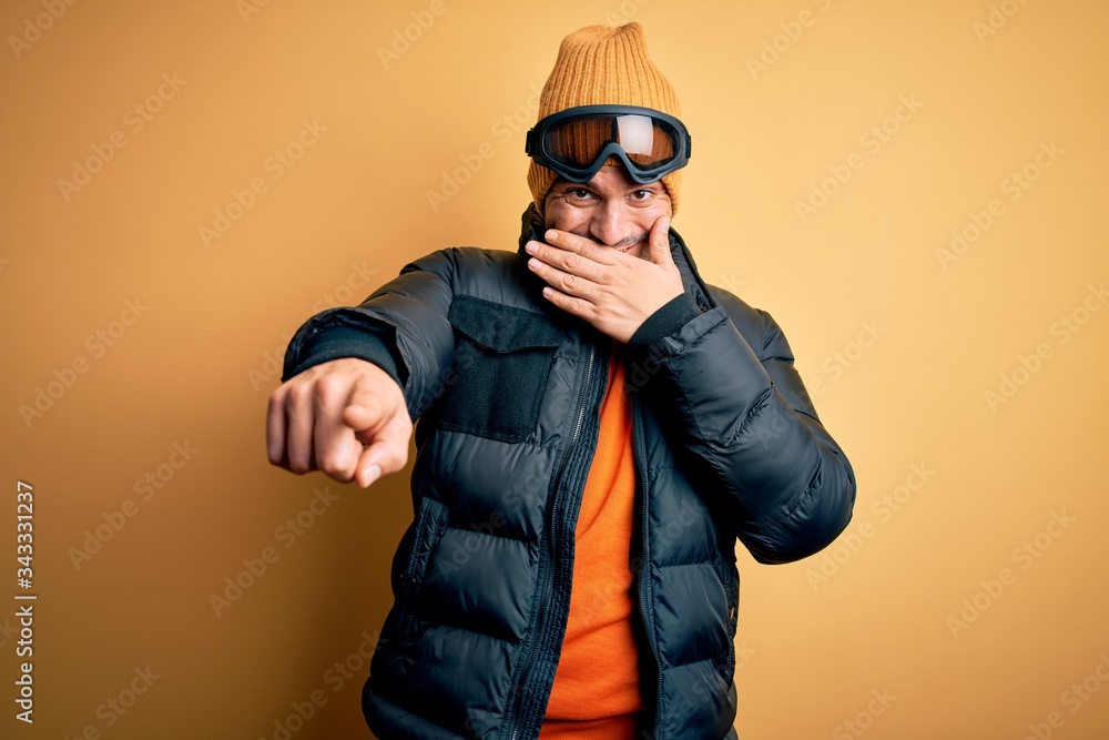 Young handsome skier man skiing wearing snow sportswear using ski goggles laughing at you, pointing finger to the camera with hand over mouth, shame expression Young handsome skier man skiing wearing snow sportswear using ski goggles laughing at you, pointing finger to the camera with hand over mouth, shame expression