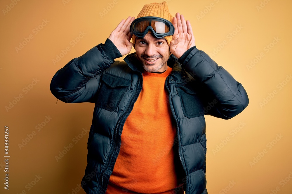 Young handsome skier man skiing wearing snow sportswear using ski goggles Trying to hear both hands on ear gesture, curious for gossip. Hearing problem, deaf Young handsome skier man skiing wearing snow sportswear using ski goggles Trying to hear both hands on ear gesture, curious for gossip. Hearing problem, deaf