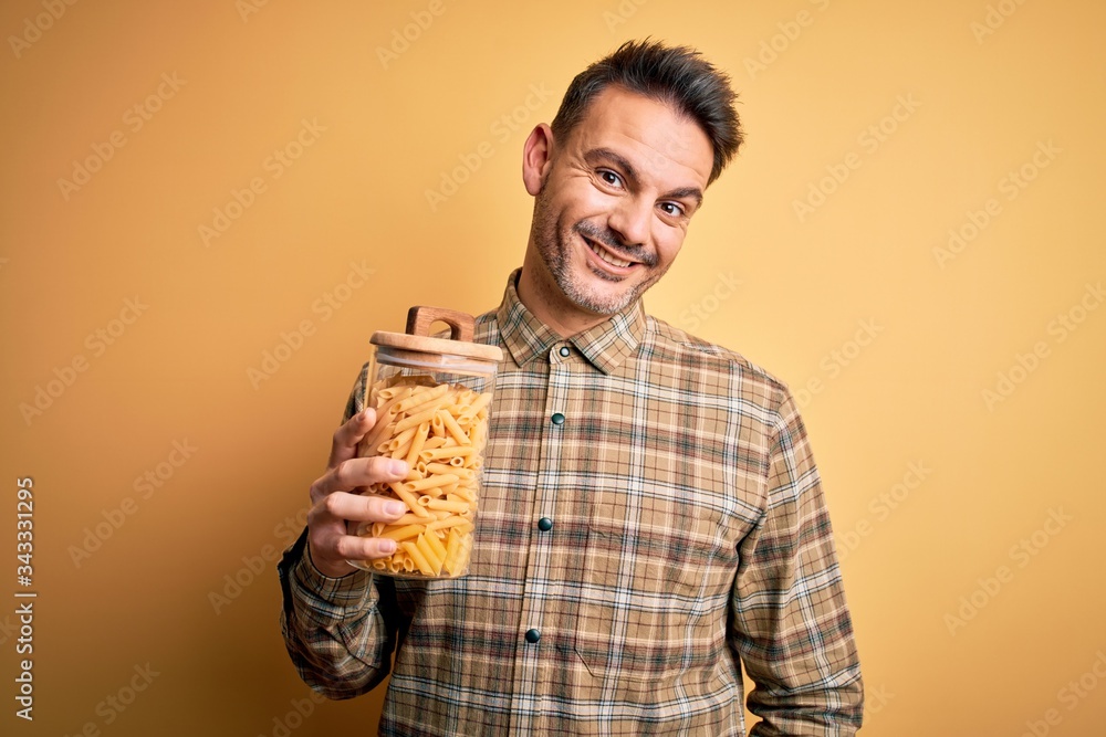 Young handsome man holding jar with dry italian pasta macaroni over yellow background with a happy face standing and smiling with a confident smile showing teeth Young handsome man holding jar with dry italian pasta macaroni over yellow background with a happy face standing and smiling with a confident smile showing teeth