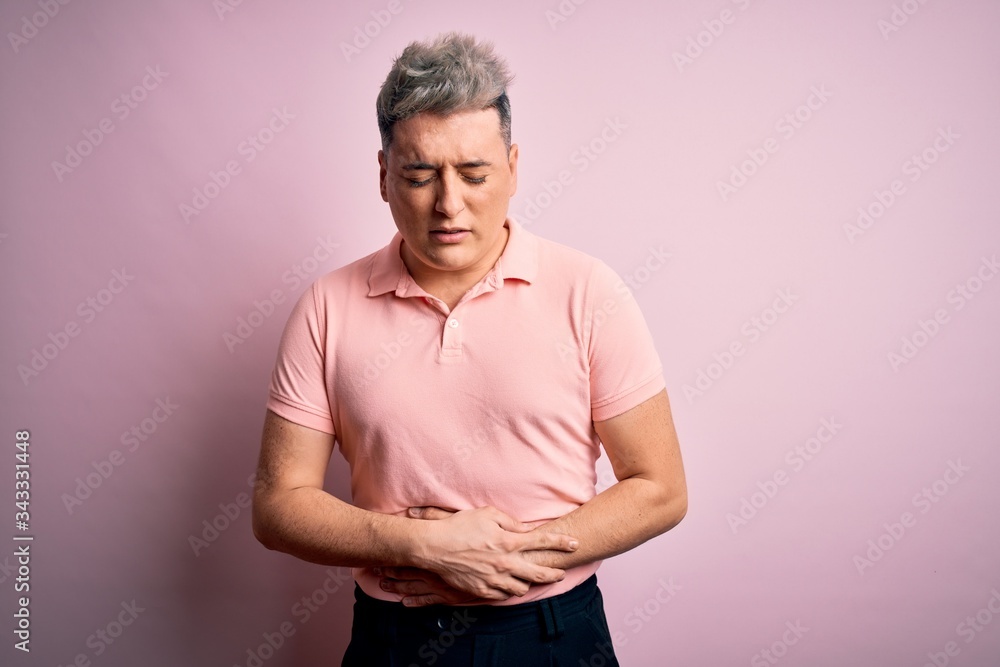 Young handsome modern man wearing casual pink t-shirt over isolated background with hand on stomach because nausea, painful disease feeling unwell. Ache concept. Young handsome modern man wearing casual pink t-shirt over isolated background with hand on stomach because nausea, painful disease feeling unwell. Ache concept.