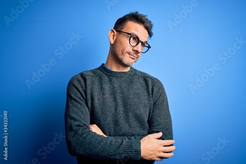 Young handsome man wearing casual sweater and glasses standing over blue background looking away to side with smile on face, natural expression. Laughing confident.
