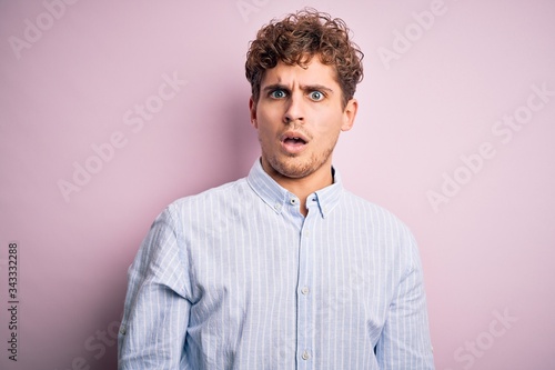 Young blond handsome man with curly hair wearing striped shirt over white background In shock face, looking skeptical and sarcastic, surprised with open mouth