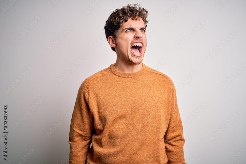 Fototapeta premium Young blond handsome man with curly hair wearing casual sweater over white background angry and mad screaming frustrated and furious, shouting with anger. Rage and aggressive concept.
