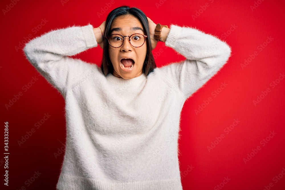 Young beautiful asian woman wearing casual sweater and glasses over red background Crazy and scared with hands on head, afraid and surprised of shock with open mouth Young beautiful asian woman wearing casual sweater and glasses over red background Crazy and scared with hands on head, afraid and surprised of shock with open mouth