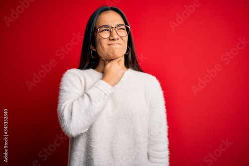 Young beautiful asian woman wearing casual sweater and glasses over red background Touching painful neck, sore throat for flu, clod and infection © Krakenimages.com