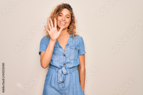 Young beautiful woman with blue eyes wearing casual denim dress over white background showing and pointing up with fingers number five while smiling confident and happy.