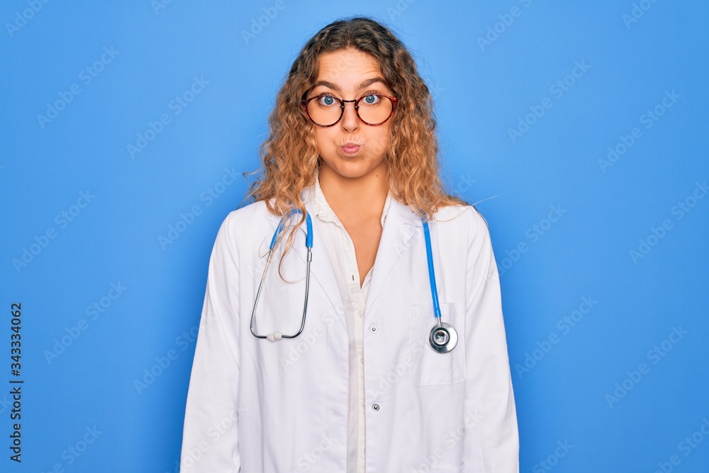 Young beautiful blonde doctor woman with blue eyes wearing coat and stethoscope puffing cheeks with funny face. Mouth inflated with air, crazy expression. Young beautiful blonde doctor woman with blue eyes wearing coat and stethoscope puffing cheeks with funny face. Mouth inflated with air, crazy expression.